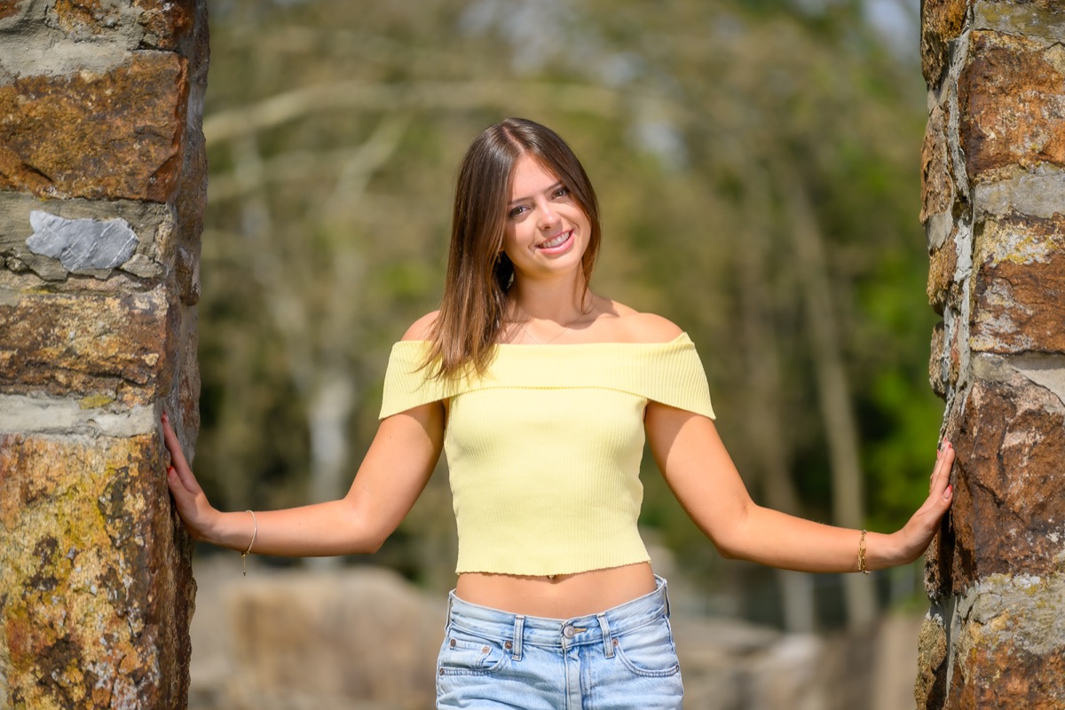 Senior portrait between stone pillars in a yellow top and jeans